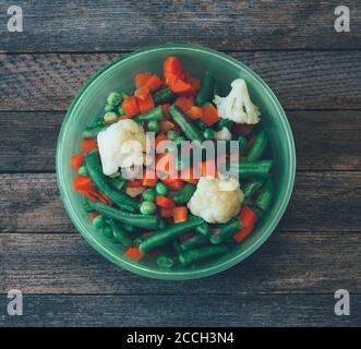 Piatti della cucina asiatica. Miscela vegetale di carote, piselli, fagioli verdi e cavolfiore in una ciotola verde sul vecchio tavolo di legno, vista ravvicinata. Pho tinta Foto Stock