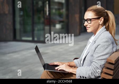 Lavorare online. Vista laterale di una donna d'affari di successo che lavora su un computer portatile mentre si siede sulla panchina all'aperto vicino all'edificio dell'ufficio Foto Stock