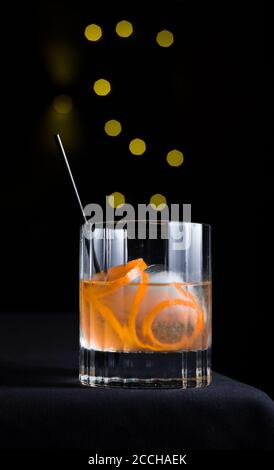 Un cocktail artigianale con guarnitura in crosta d'arancia servito sulla rocce isolate su sfondo nero con cerchi di bokeh gialli Foto Stock