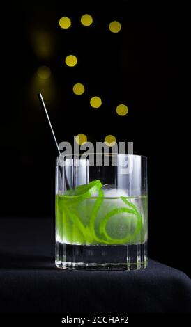 Vista isolata di un cocktail artigianale con guarnitura di crosta di lime servito in un vetro di rocce su uno sfondo nero con bokeh giallo Foto Stock