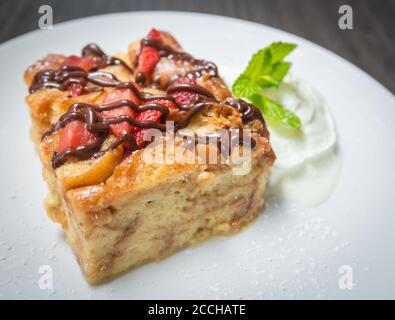 Un'ampia fetta di budino di pane fatto in casa condito con fragole fresche e cioccolato, servito con panna montata e menta Foto Stock