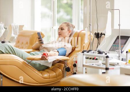 Giovane donna donatrice sdraiata sul divano e che la usa telefono cellulare durante la donazione di sangue in ospedale Foto Stock