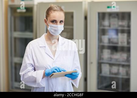 Ritratto di giovane dottoressa in maschera e in bianco cappotto guardando la fotocamera mentre si utilizza il tablet digitale in ospedale Foto Stock