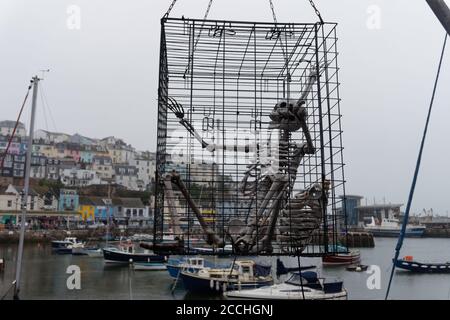 Scheletro di faux in una grande gabbia di filo sospeso sul porto di Brixham. Barche ormeggiate nel porto e case sul molo. Foto Stock