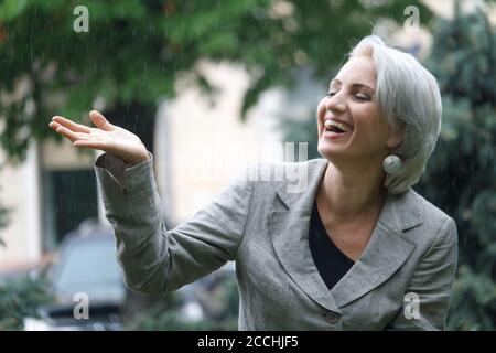 Donna bionda matura felice cammina in pioggia e cattura gocce con mano, fuoco selettivo Foto Stock