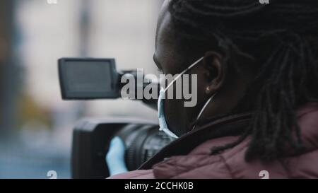 Primo piano, videografo nero africano con maschera mediale che guarda attraverso il mirino sulla videocamera . Foto di alta qualità Foto Stock