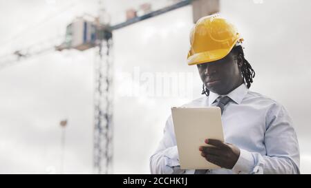 Ritratto di un uomo africano con hardhat utilizzando la tavoletta sul cantiere e osservando il lavoro. Foto di alta qualità Foto Stock