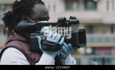 Primo piano, videografo nero africano con maschera mediale che guarda attraverso il mirino sulla videocamera . Foto di alta qualità Foto Stock