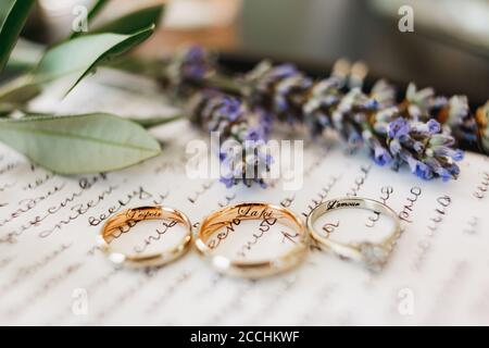 Primo piano degli anelli di nozze della sposa e dello sposo e degli anelli d'oro bianchi con iscrizioni su un pezzo di carta con un giuramento e uno sprig di lavanda. Foto Stock
