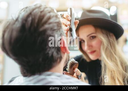 Donna barbiere taglio e taglio barba di cliente nel suo negozio Foto Stock