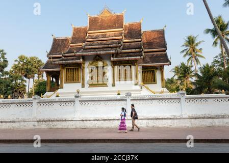 Persone che camminano per strada a Luang Prabang con Haw Pha Bang sullo sfondo Foto Stock