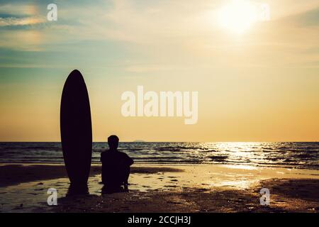 Silhouette di surf uomo seduto con una tavola da surf sulla spiaggia di mare al tramonto. Bel modello di uomo asiatico nei suoi anni 20. Foto Stock