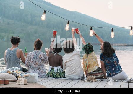 Vista posteriore del gruppo di amici che tostano con bicchieri di birra e celebrare la loro vacanza estiva mentre si siede su un molo Foto Stock