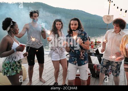 Gruppo di giovani con bevande e sparklers che ballano un molo durante la festa Foto Stock