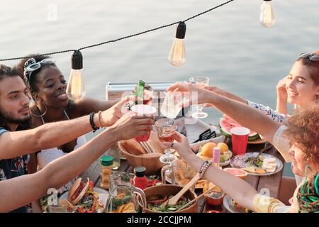 Gruppo di amici seduti al tavolo da pranzo e tostatura con cocktail che celebrano le vacanze all'aperto su un molo Foto Stock