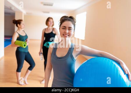 Una giovane donna con una palla ginnica in mano sorridente e in posa per la macchina fotografica. Sullo sfondo ci sono due donne con tappetini nelle loro mani. Th Foto Stock