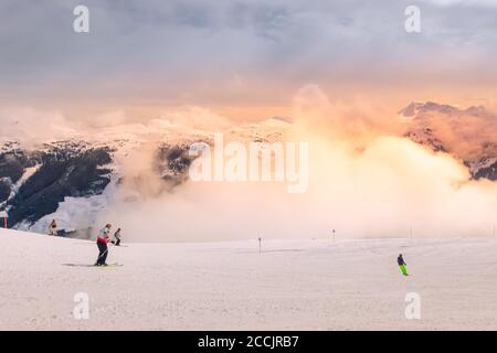 Saalbach, Austria - 6 marzo 2020: Sciatori e snowboarder sulle piste da sci in località invernale di autestrain, cime innevate delle Alpi e nuvole serali Foto Stock