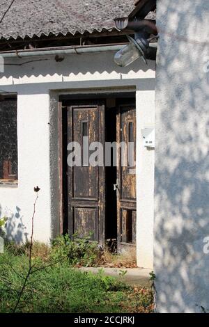 Ingresso angolo alla vecchia casa suburbana di famiglia abbandonata in forma di porte in legno dilatate parzialmente aperte con vernice incrinata montata sulla facciata bianca Foto Stock