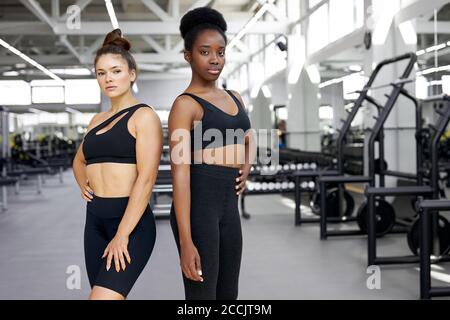ritratto di due diverse ragazze sportive in palestra, giovani donne africane e caucasiche in abbigliamento sportivo guardare la macchina fotografica, dopo esercizi di crossfit Foto Stock