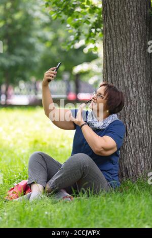 Donna anziana nel parco centrale della città che ha una chiamata online con il suo smartphone. Lei dice ciao con palmo di mano. Foto Stock