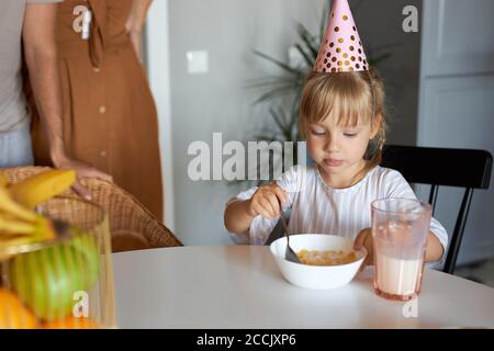 bella adorabile ragazza seduta in cucina da sola, in cappello di festa, prima di festa di compleanno a casa. mangiare, mangiare al mattino Foto Stock