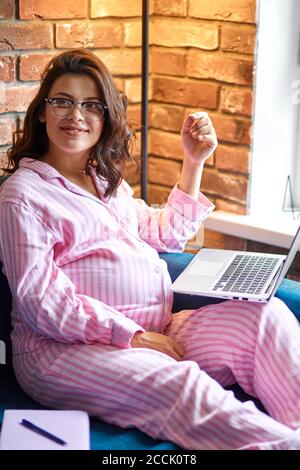 adorabile donna incinta pacifica che lavora con laptop, indossando abiti casalinga. bella brunetta con pancia rotonda su freelance Foto Stock