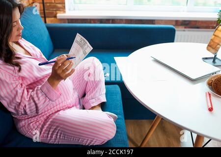soddisfatta giovane donna caucasica incinta che legge il suo calendario di gravidanza, sedersi sul divano in abbigliamento domestico Foto Stock