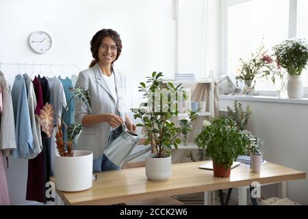 Sorridente giovane donna annaffiatura impianti in moderno ufficio creativo. Foto Stock