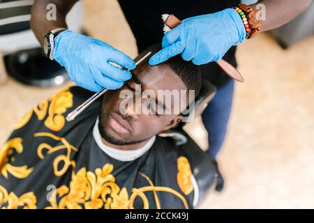 Primo piano del barbiere che indossa guanti che tagliano i capelli del cliente con il rasoio lama in salone Foto Stock