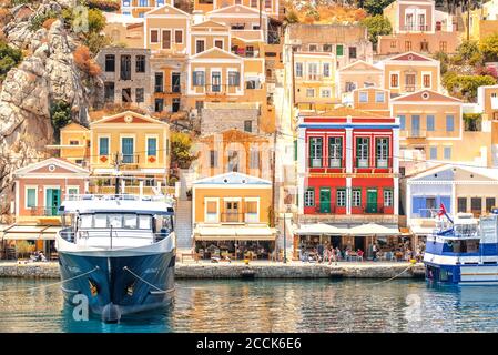 SYMI, Grecia - 27 giugno 2019: Vista della costa dell'isola di Symi con la città con case multicolore, alberi e popoli e baia con le barche in estate giorno Foto Stock