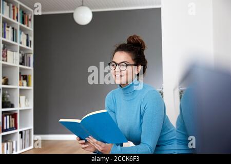 Donna premurosa che guarda via mentre tiene il libro nel soggiorno a casa Foto Stock