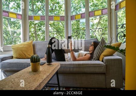 Donna che legge libro mentre si rilassa sul divano in soggiorno Foto Stock