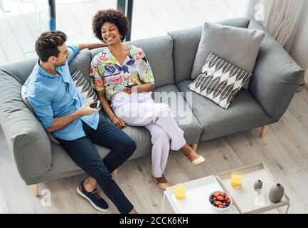 Felice coppia multietnica che tiene smartphone mentre si siede sul divano in attico