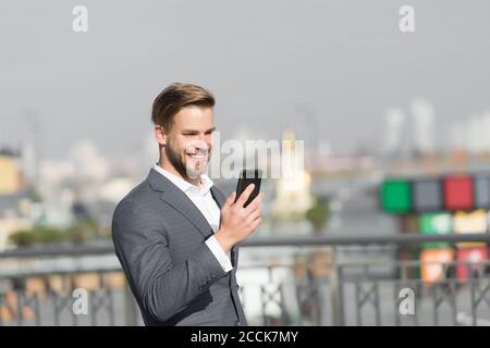 Imprenditore sorridenti utilizza lo smartphone per le video chiamate o messaggi di testo, skyline background. Business il concetto di chiamata. Uomo in tuta imprenditore assume i vantaggi delle moderne tecnologie mobili per il business. Foto Stock