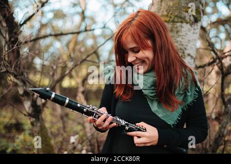 Felice giovane donna che guarda il clarinetto mentre si trova contro l'albero tronco in foresta Foto Stock