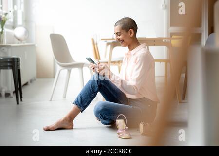 Donna felice seduta sul pavimento in un loft usando smartphone Foto Stock