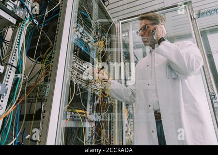 Uomo maturo che parla con lo smartphone mentre si collega il ricetrasmettitore cavo in fibra ottica nel data center Foto Stock