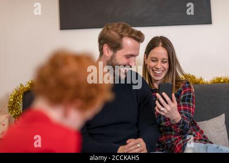 I genitori allegri videochiamano con lo smartphone mentre sono seduti divano a casa durante il Natale Foto Stock