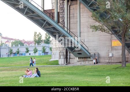 Novosibirsk, Russia - 07.12.2020: Giovani ragazze si siedono sull'erba verde durante un picnic sotto un grande ponte di ferro sull'argine. Foto Stock