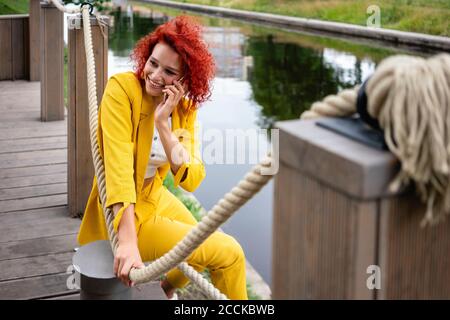 Giovane donna con capelli ricci e tuta gialla seduta sul lungofiume, che parla al telefono Foto Stock