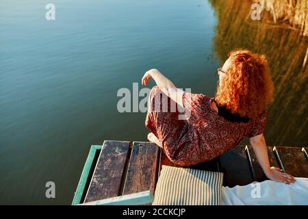 Giovane donna decapitata seduta sul molo presso un lago a. tramonto Foto Stock