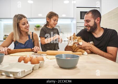 Madre impastare l'impasto con il mattarello mentre padre e figlia giocare con il gatto sul tavolo della cucina Foto Stock