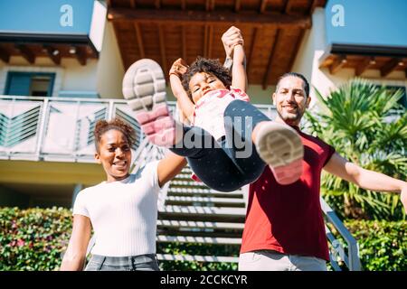 Felici genitori che tengono le mani giocose della figlia e la raccolgono contro casa Foto Stock