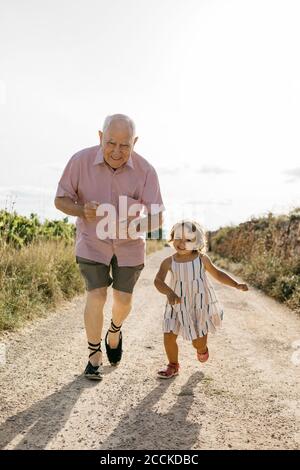 Uomo anziano giocoso che corre con la nipote sulla strada sterrata in mezzo piante durante la giornata di sole Foto Stock