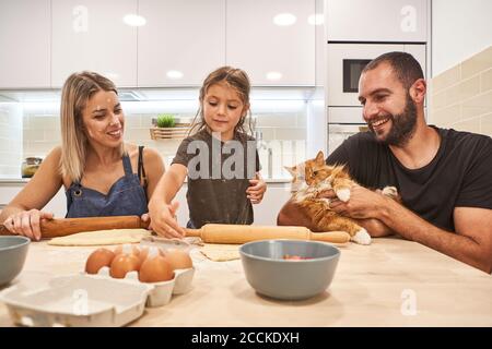 Madre e figlia impastano l'impasto con il mattarello mentre padre tenendo gatto sul tavolo della cucina Foto Stock