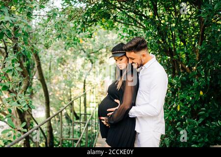 Uomo sorridente che abbraccia e tocca il ventre della donna mentre si è in piedi nel parco Foto Stock