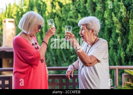 Le amiche senior tostano flauti di champagne mentre si levano in piedi dalla ringhiera in balcone Foto Stock