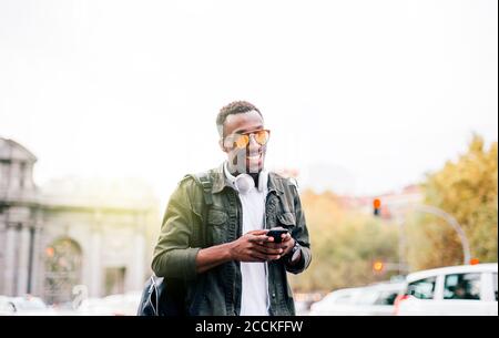 Uomo sorridente che indossa occhiali da sole utilizzando uno smartphone mentre si sta in piedi cielo limpido in città Foto Stock