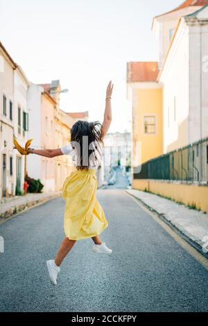 Donna spensierata con capelli lunghi che saltano sulla strada tra gli edifici in città Foto Stock