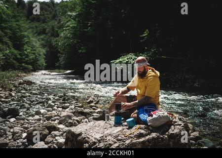 Escursionista con barba completa e felpa con cappuccio gialla durante la pausa, cucinando il tè sul lungofiume Foto Stock